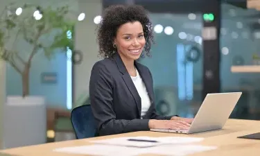 A professional woman sits at a desk with a laptop, smiling confidently, representing career success with a business management degree.